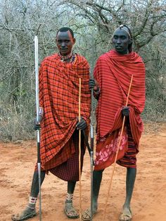 two men in red and black outfits with sticks standing on dirt ground next to trees