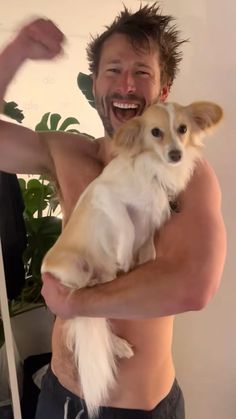 a man holding a dog in his arms and smiling at the camera while standing next to a potted plant