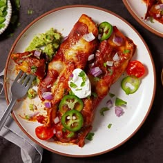 two white plates topped with enchiladas and vegetables next to a fork on a table