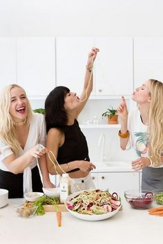 three women are laughing while preparing food in the kitchen, one woman is holding a string above her head