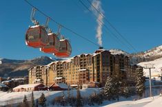 a ski lift with two people on it above the snow covered ground and buildings in the background