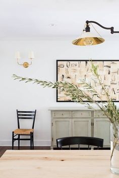 a vase with some plants in it sitting on a table next to two chairs and a painting