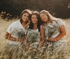 three women are sitting in the tall grass with baby's breath flowers on their hands