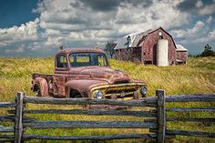 an old rusted truck is parked in the grass near a wooden fence and barn