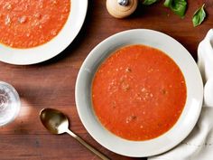 two bowls of tomato soup on a wooden table with spoons and silverware next to it