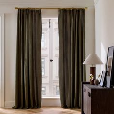 a living room with a large window covered in curtains next to a wooden table and lamp