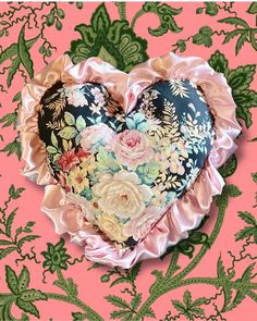 a heart shaped pillow sitting on top of a pink wallpaper covered in green leaves and flowers