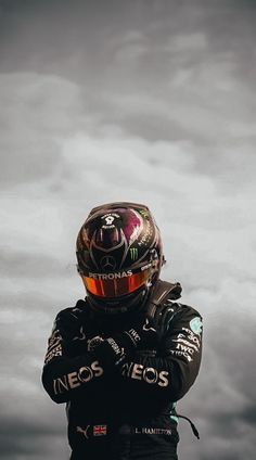 a man wearing a racing suit and helmet standing in front of a cloudy sky with his hands on his hips