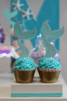 two cupcakes with blue frosting and a mermaid tail on top are sitting on a table