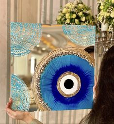 a woman holding up a blue and white decorative piece in front of a mirror with an eye on it