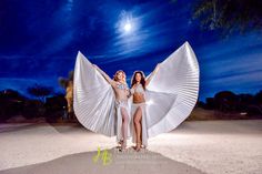 two women in bikinis standing on the beach with their arms around each other and holding large white wings