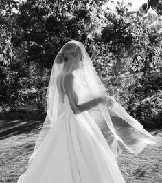 a woman in a wedding dress and veil walking through the grass with trees behind her