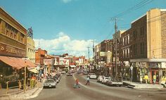 an old photo of people crossing the street in front of stores and cars on a sunny day