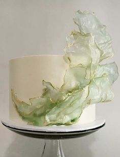 a white and green wedding cake with gold leaf decoration on top, sitting on a glass stand