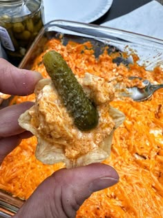 a hand holding a cracker with a pickle on it in front of a casserole dish