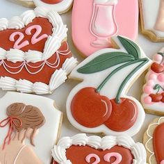 many decorated cookies are displayed on a white tablecloth with pink and red accents, including cherries