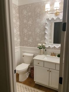 a white toilet sitting under a bathroom mirror next to a sink and a vanity with flowers on it