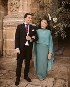 a man and woman standing next to each other in front of a stone building with an olive tree