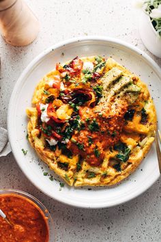 a white plate topped with an omelet covered in toppings next to a bowl of sauce