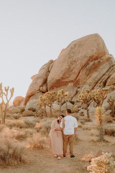 a man and woman standing in the desert with joshua trees behind them, looking at each other