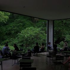 people are sitting at tables in an open room with large windows looking out onto the woods