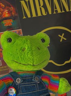 a green stuffed animal with overalls and a rainbow striped shirt is posed in front of a movie poster