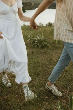 a man and woman holding hands while walking through the grass near a body of water