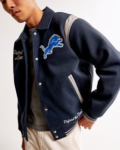 Classic bomber jacket in a soft faux wool fabric and relaxed-fit silhouette, featuring Detroit Lions varsity-inspired embroidered graphic patch details throughout, side pockets, snap-up front and tipping details at elastic cuffs and waistband. Fitted Varsity Outerwear For Work, Fall Varsity Sport Coat With Ribbed Cuffs, Varsity Sport Coat With Ribbed Cuffs For Fall, Winter Varsity Style Outerwear For Work, Collegiate Style Winter Fleece Outerwear, Collegiate Outerwear With Pockets, Collegiate Outerwear With Pockets For Fall, Fitted Varsity Outerwear For Fall, Navy Varsity Outerwear For Fall