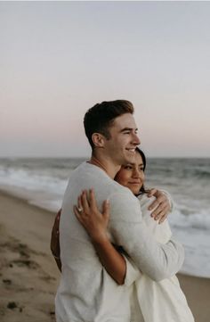 a man and woman hugging on the beach at sunset with waves crashing in behind them