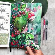 a hand holding an open notebook with a green bird on it and red berries in the background