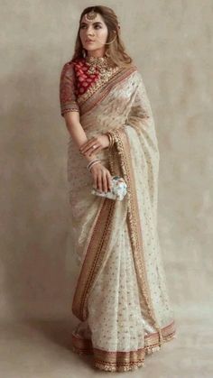 a woman wearing a white sari with red and gold accents on her chest, standing in front of a beige wall