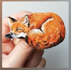 a hand holding a miniature fox brooch in it's right hand, with the image of a sleeping fox on its left side