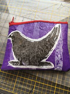 a purple bag with a black bird on it sitting on top of a piece of fabric