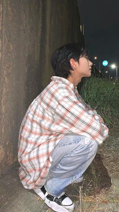 a young man squatting on the ground next to a cement wall at night with street lights in the background