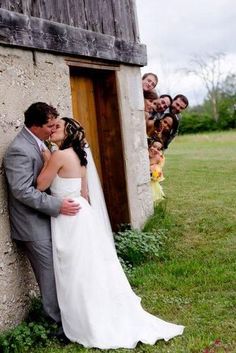 a bride and groom kissing in front of an outhouse with their wedding party behind them