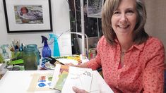 a woman sitting at a desk holding an open book with pictures on the wall behind her