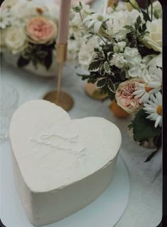 a white heart shaped cake sitting on top of a table next to flowers and candles