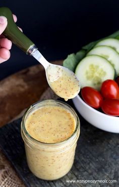 a person holding a spoon over a jar filled with dressing next to a bowl of vegetables and cucumbers