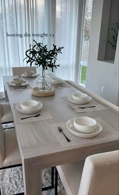 a dining room table with white plates and place settings on it, in front of a window