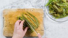 a person cutting up some green beans on a wooden board next to a glass bowl