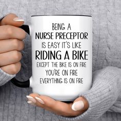 a woman holding a coffee mug with the words being a nurse preceptor is easy it's like riding a bike except the bike on fire you're on fire