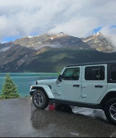 a white jeep parked on the side of a road next to a mountain lake with snow capped mountains in the background