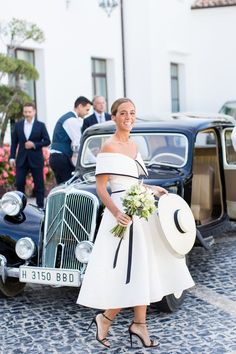 a woman in a white dress and hat standing next to an old fashioned black car