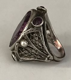 "This lovely filigreed and ornate ring has an oval shaped faceted amethyst purple color glass stone at its center. It is flanked on either side of to and bottom with small round bezel set purple glass stones. The sides have lattice shaped cut outs and the band is smooth. The face of this ring is about 3/4\" long and about 1/2\" across. It is a size 3 1/4 and is marked Sterling. Please note there are some overall signs of wear, especially to the stones, four of five of which, including the center Formal Purple Oval Ruby Ring, Heirloom Amethyst Filigree Ring In Purple, Heirloom Amethyst Ring With Filigree, Heirloom Purple Amethyst Ring With Intricate Design, Heirloom Style Purple Amethyst Ring With Filigree, Antique Oval Amethyst Ring Collectible, Antique Purple Amethyst Ring With Intricate Design, Purple Amethyst Rings With Intricate Design, Heirloom Style Purple Oval Amethyst Ring