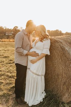 a pregnant couple kissing in front of a hay bale at sunset with the sun shining on them