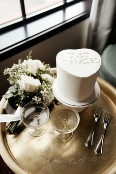 a table topped with a white cake next to two wine glasses and silverware on top of a gold tray