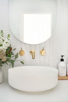 a white sink sitting under a round mirror next to a vase with flowers in it