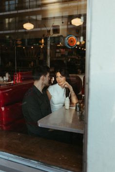 a man and woman sitting at a table in front of a restaurant window talking on their cell phones