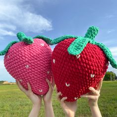 two crocheted strawberries being held up in the air by their hands, against a blue sky