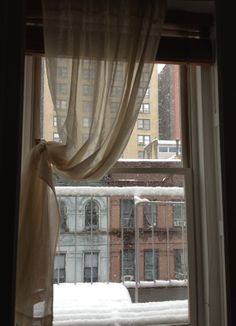 a window with curtains and snow on the ground in front of it, looking out at buildings